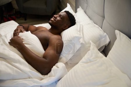 Téléchargez les photos : Souriant mec avec un torse nu jouit de détente matinale dans un lit doux, il est assis sur des oreillers doux - en image libre de droit