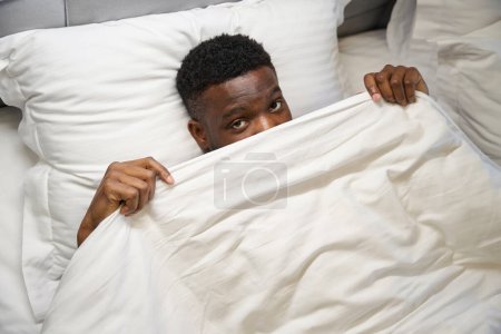 Photo for African American man looks out from under the blanket as, he lies on a soft bed - Royalty Free Image