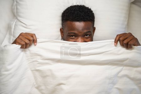 Photo for Happy African American man looks out from under the blanket as, he lies on a soft bed - Royalty Free Image