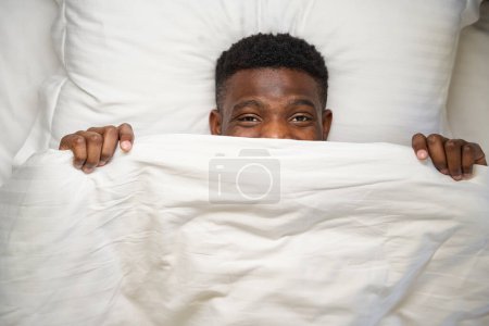 Photo for Happy African American male looks out from under the blanket, he lies on a soft bed - Royalty Free Image