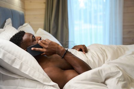 Téléchargez les photos : Homme aux cheveux bouclés communique sur un téléphone portable au lit, il se trouve sous une couverture douce - en image libre de droit