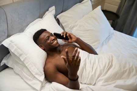 Téléchargez les photos : Afro-Américain avec un torse nu communique sur un téléphone portable, il s'allonge sur un lit doux - en image libre de droit
