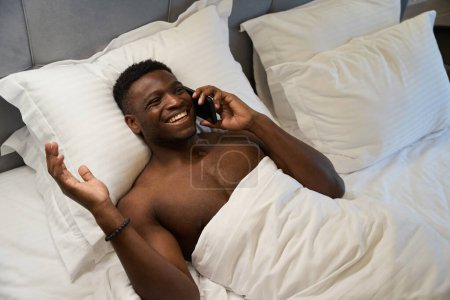 Photo for African American male with a naked torso communicates on a mobile phone, he lies on a soft bed - Royalty Free Image