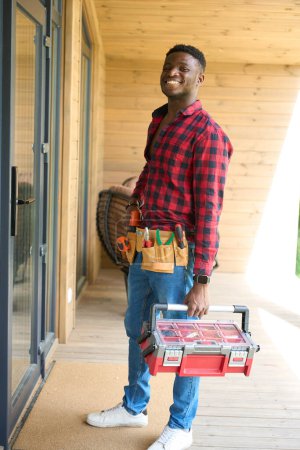 Photo for Smiling guy stands at the front door with a toolbox, he is wearing a red plaid shirt and jeans - Royalty Free Image