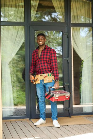 Foto de Hombre sonriente se para en una terraza de madera con una caja de herramientas, lleva una camisa roja a cuadros y vaqueros - Imagen libre de derechos