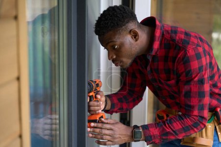Photo for African American guy is repairing a glass door, he is wearing a red plaid shirt and jeans - Royalty Free Image