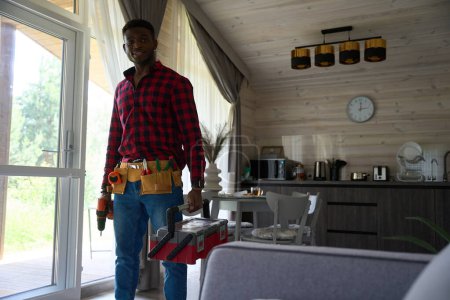 Téléchargez les photos : Travailleur afro-américain dans une pièce lumineuse avec des outils et une perceuse, un gars en chemise à carreaux - en image libre de droit