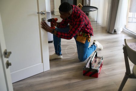 Photo for African American housekeeper repairs a wooden door, he uses a drill - Royalty Free Image