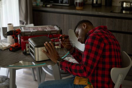 Photo for Young repairman is repairing a toaster on the kitchen table, he is using a screwdriver - Royalty Free Image