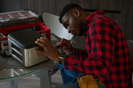 Foto de Reparador de pelo rizado está reparando una tostadora en la mesa de la cocina, utiliza un destornillador - Imagen libre de derechos