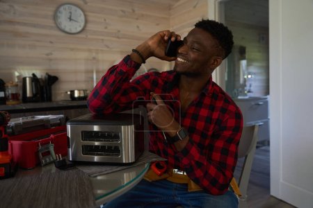 Téléchargez les photos : Un homme souriant communique sur un téléphone portable, il fait des réparations mineures dans la cuisine - en image libre de droit
