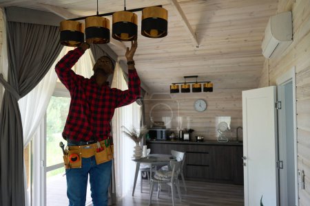 Photo for African American guy is repairing a chandelier in the kitchen, he has a tool belt - Royalty Free Image