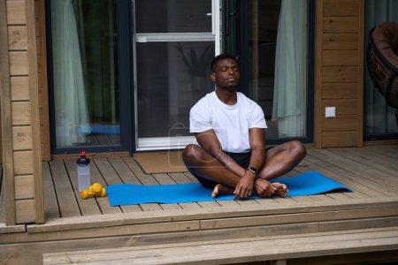 Photo for African American guy practices yoga on a wooden terrace, he sits on a karimat - Royalty Free Image