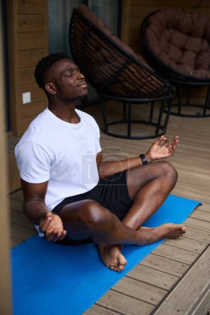 Photo for African American male practices yoga on a wooden terrace, he meditates in the lotus position - Royalty Free Image