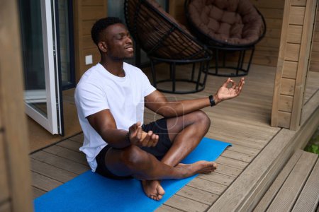 Foto de Sonriente hombre practica yoga en una terraza de madera, medita en la posición de loto - Imagen libre de derechos