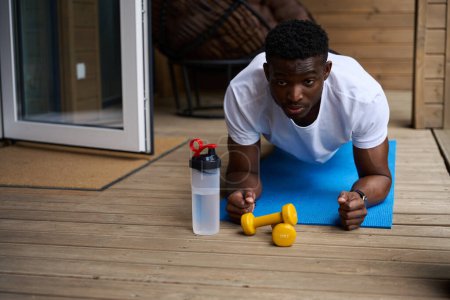 Photo for Male in sportswear doing fitness on the terrace of an eco-cottage, next to dumbbells and a bottle of water - Royalty Free Image