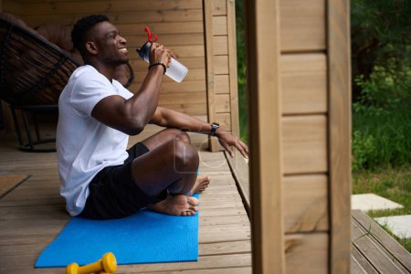 Téléchargez les photos : Afro-Américain boit de l'eau pendant une pause entre les exercices, il est situé sur le karimat - en image libre de droit