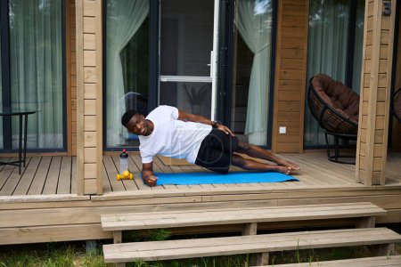 Téléchargez les photos : Jeune homme en tenue de sport fait du fitness sur la terrasse de l'éco-chalet, à côté des haltères et une bouteille d'eau - en image libre de droit
