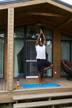 Photo for Young male in sportswear performs asana on the terrace of an eco-cottage, next to dumbbells and a bottle of water - Royalty Free Image