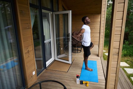 Photo for Man in sportswear performs an asana on the terrace of an eco-cottage, with dumbbells and a bottle of water nearby - Royalty Free Image