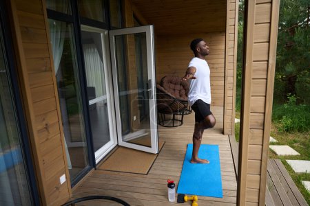 Photo for Guy in sportswear performs an asana on the terrace of an eco-cottage, next to dumbbells and a bottle of water - Royalty Free Image
