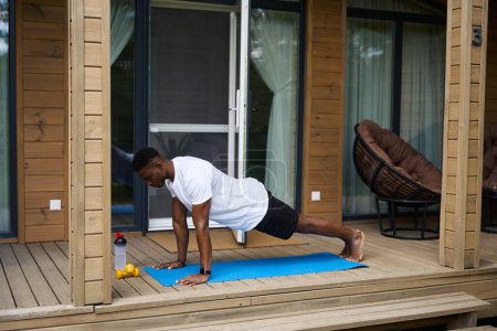 Téléchargez les photos : Afro-Américain faisant de la remise en forme sur la terrasse d'un éco-chalet, à côté d'haltères et d'une bouteille d'eau - en image libre de droit