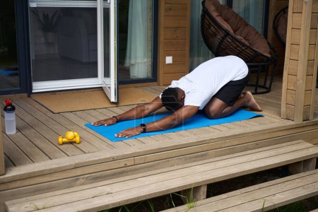 Photo for African American man doing fitness on the terrace of an eco-cottage, next to dumbbells and a bottle of water - Royalty Free Image