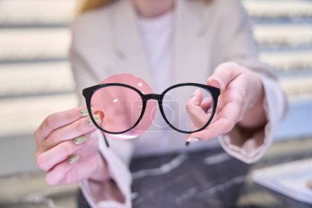 Foto de Mujer sostiene gafas y un aparato oftálmico en sus manos, un especialista en su lugar de trabajo en un salón de óptica - Imagen libre de derechos