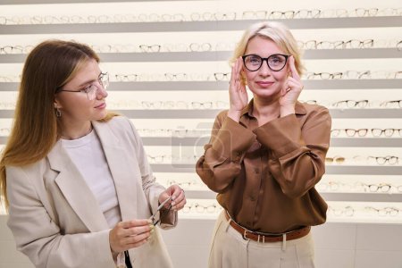 Téléchargez les photos : Un client d'âge moyen essaie des lunettes dans un salon d'optique, avec un consultant en ophtalmologie à proximité - en image libre de droit