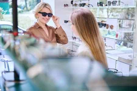 Photo for Charming blonde woman trying on sunglasses in an optics salon, next to an ophthalmologist consultant - Royalty Free Image
