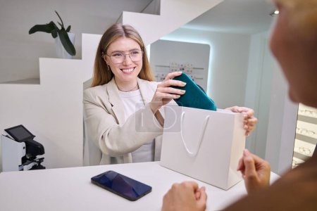 Photo for Optical salon consultant puts a case with glasses in a gift bag, a mobile phone lies on the counter - Royalty Free Image