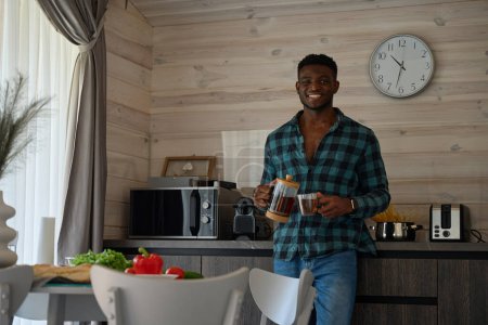 Foto de El hombre afroamericano se sienta en una acogedora cocina, vierte té en una taza - Imagen libre de derechos