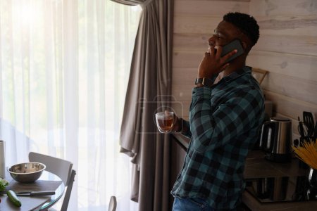Téléchargez les photos : Le gars communique en ligne sur son téléphone portable, il s'assoit avec une tasse de thé dans la cuisine - en image libre de droit