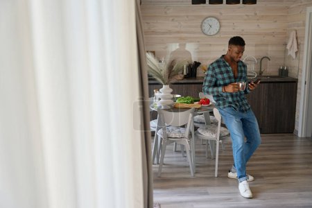 Photo for African American guy communicates online on his mobile phone, he sits down with cup of tea in the kitchen area - Royalty Free Image