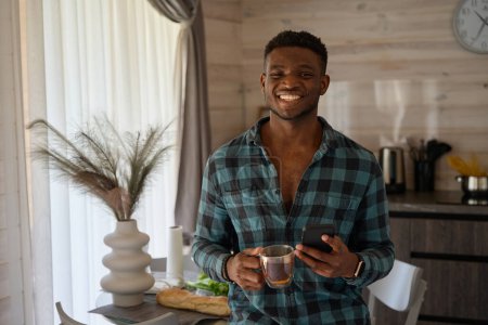 Foto de El tipo feliz está sentado con una taza de té en una acogedora cocina, tiene un teléfono móvil en la mano - Imagen libre de derechos