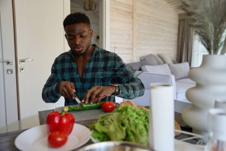 Foto de Hombre afroamericano se sienta a la mesa y corta verduras para una ensalada, él está en ropa casual cómoda - Imagen libre de derechos