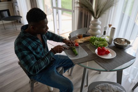 Téléchargez les photos : Homme assis à une table et côtelettes légumes pour une salade, il est dans des vêtements décontractés confortables - en image libre de droit