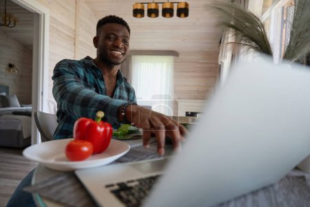 Foto de Chico afroamericano se sienta en una acogedora cocina con un ordenador portátil, verduras jugosas en la mesa - Imagen libre de derechos