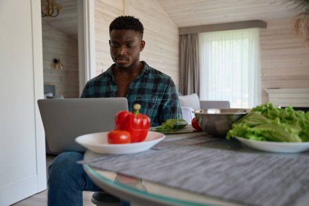 Foto de Joven se sienta con un ordenador portátil en la cocina, hay platos y comida en la mesa - Imagen libre de derechos