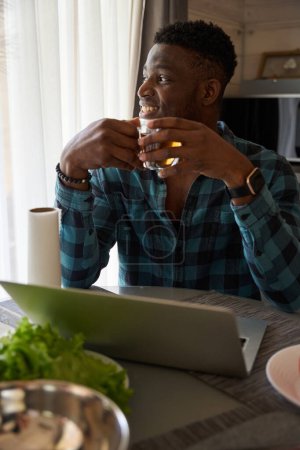 Photo for African American man with cup of tea in the kitchen with a laptop, juicy vegetables for salad on the table - Royalty Free Image