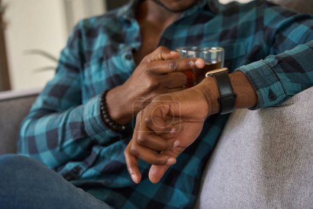 Photo for African American male sits with a cup of tea on cozy sofa, he looks at smart watch on his hand - Royalty Free Image