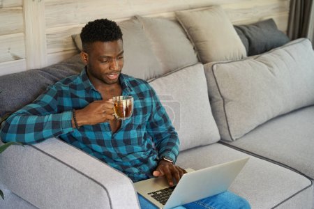 Photo for African American male sits with a cup of tea on a cozy sofa, he works online using a laptop - Royalty Free Image