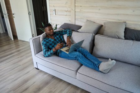 Photo for African American guy is lying with his mobile phone and laptop on the sofa, he is in comfortable home clothes - Royalty Free Image