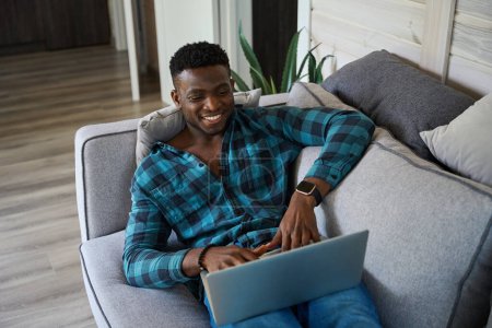 Foto de Hombre afroamericano se comunica en un ordenador portátil en el sofá, él está en ropa cómoda para el hogar - Imagen libre de derechos
