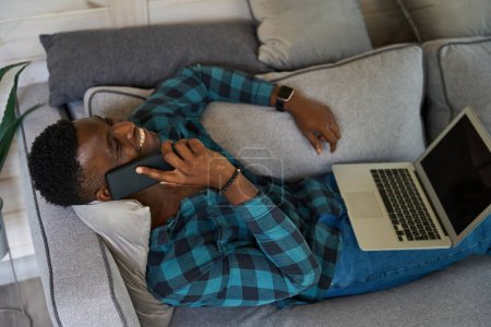 Téléchargez les photos : Homme aux cheveux bouclés assis sur un canapé confortable avec un téléphone portable et un ordinateur portable, il est dans des vêtements de maison confortables - en image libre de droit