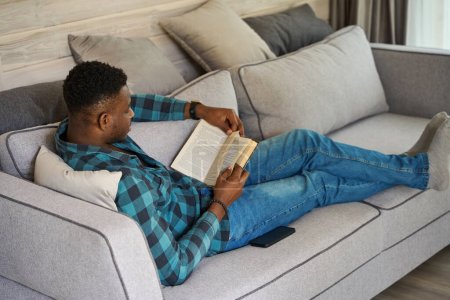 Photo for Curly-haired man in casual clothes sits comfortably on the sofa, he enjoys reading a book - Royalty Free Image