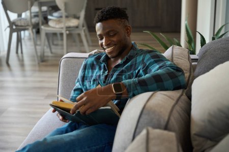 Photo for Happy African American guy sitting on the sofa, he is reading a book with pleasure - Royalty Free Image