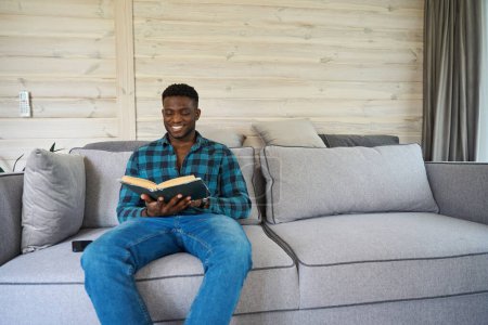 Photo for Happy African American man sitting on the sofa with a book, a mobile phone lying next to him - Royalty Free Image