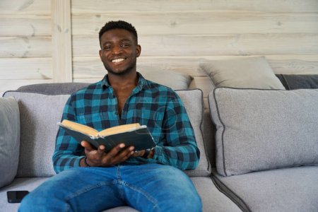 Photo for Young African American man sitting on the sofa with a book, a mobile phone lying next to him - Royalty Free Image