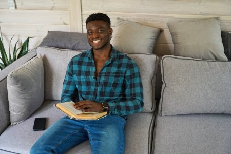 Téléchargez les photos : Mignon Afro-Américain assis sur le canapé avec un livre, un téléphone portable couché à côté de lui - en image libre de droit